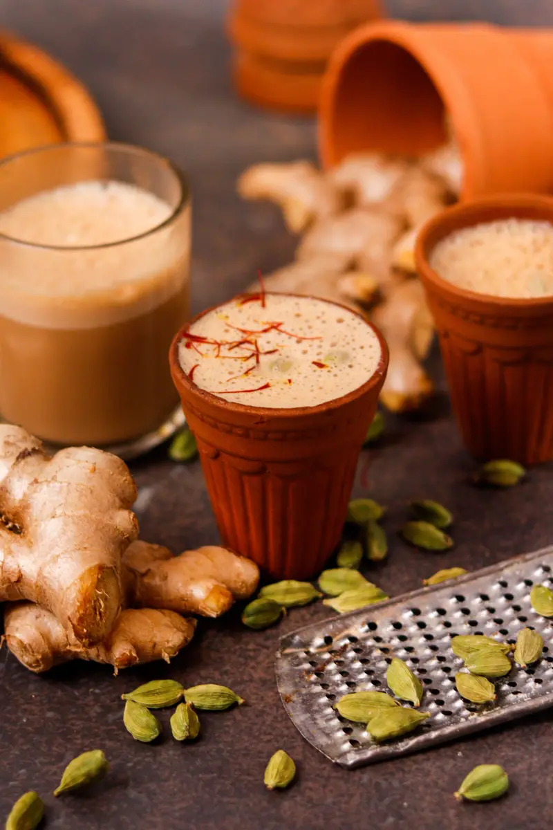 Masala chai in clay cups, topped with saffron, beside fresh ginger and green cardamom