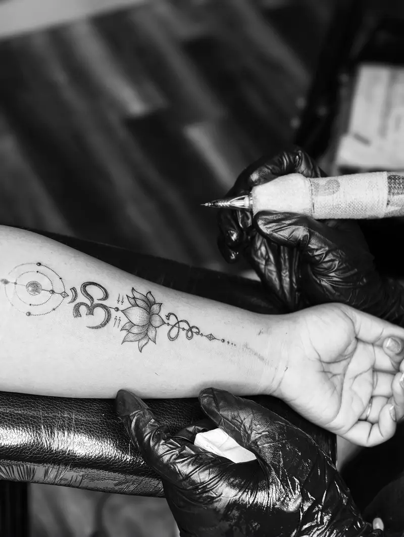 Artist in black gloves tattooing a lotus and Om line design along a forearm