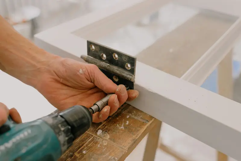 Hands holding a metal hinge against a white window frame while a cordless drill drives in a screw